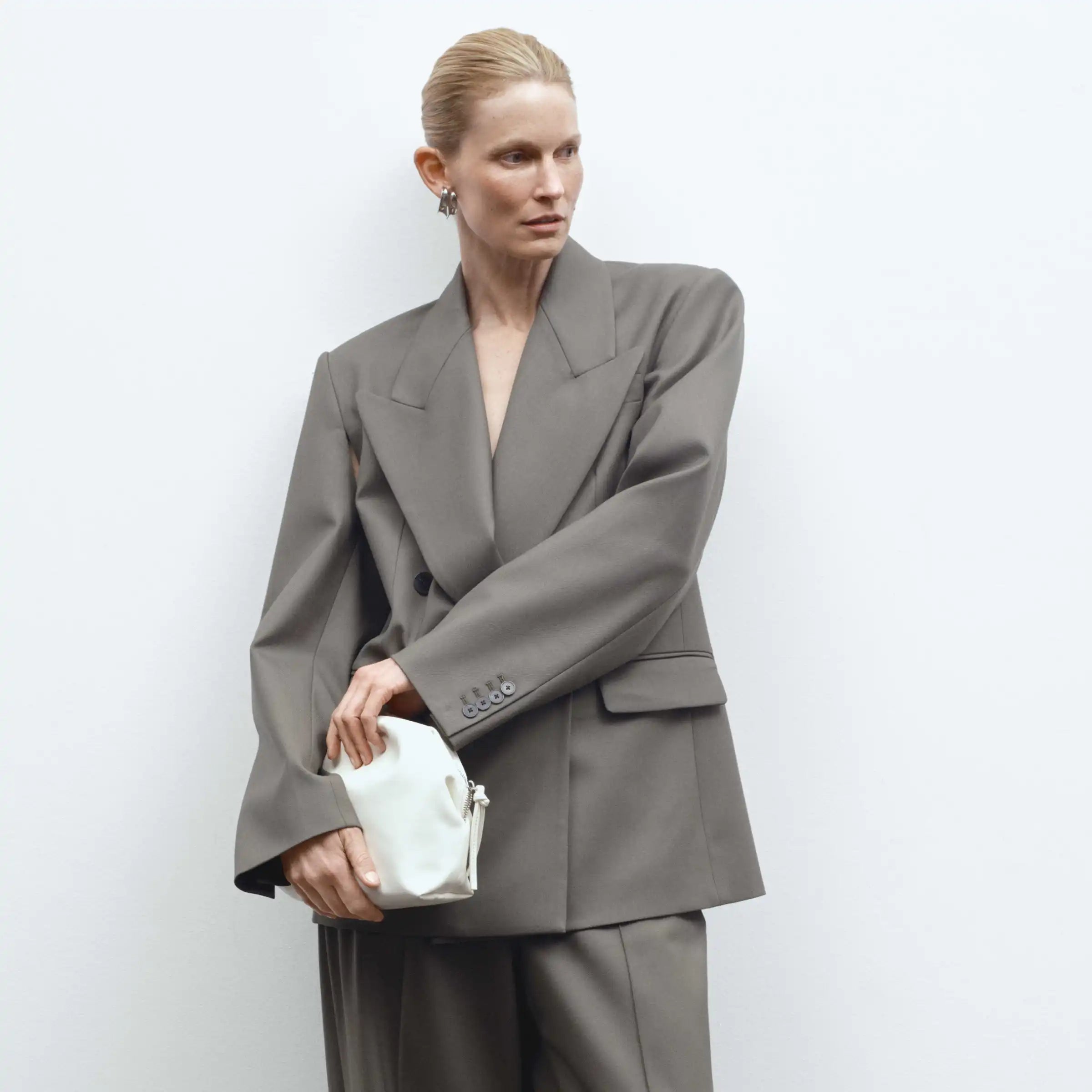 White rectangular bag with gathered sides held by woman in gray blazer and trousers.