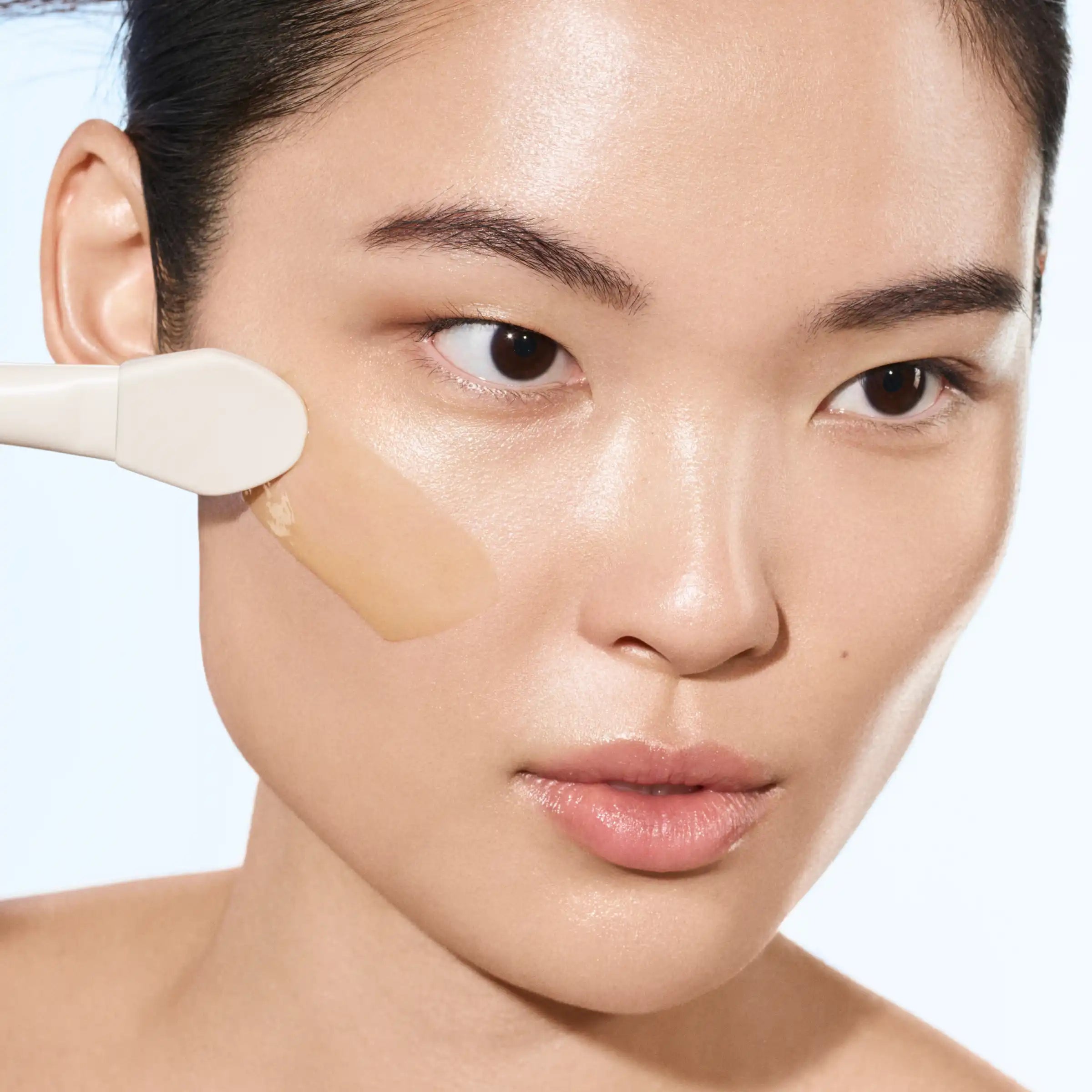 Person applying beige liquid product to cheek with white rounded brush applicator.