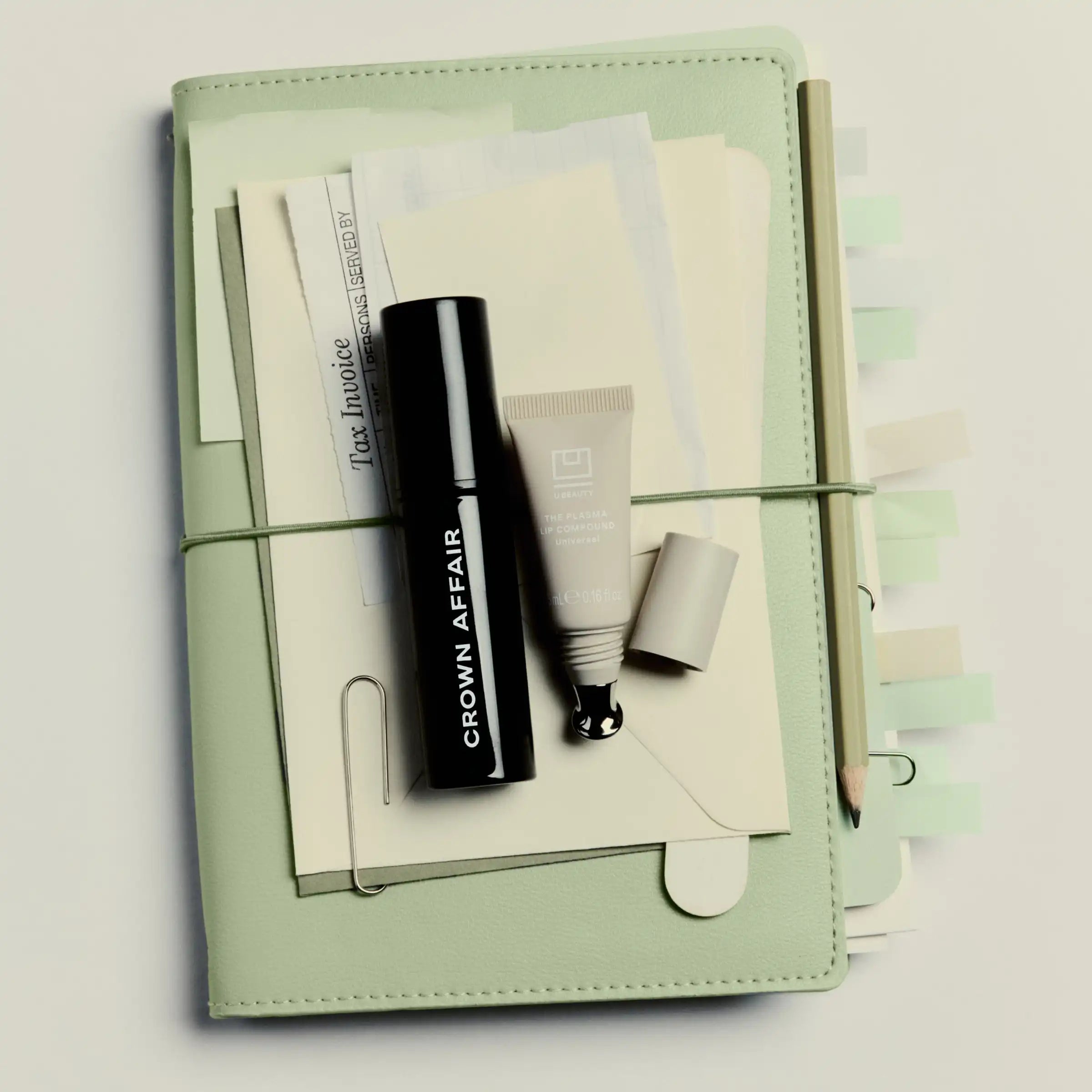 Sage green organizer containing black mascara tube, cream-colored cosmetic product, and notepad with stationery accessories.