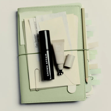 Sage green organizer containing black mascara tube, cream-colored cosmetic product, and notepad with stationery accessories.