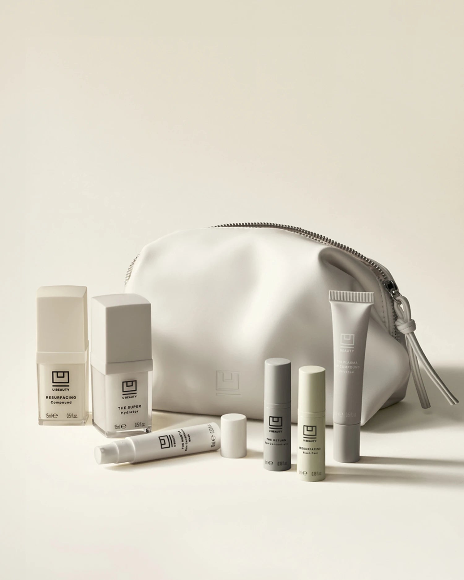 White skincare set with seven products including bottles, tubes, and a metallic silver zip pouch on beige background.
