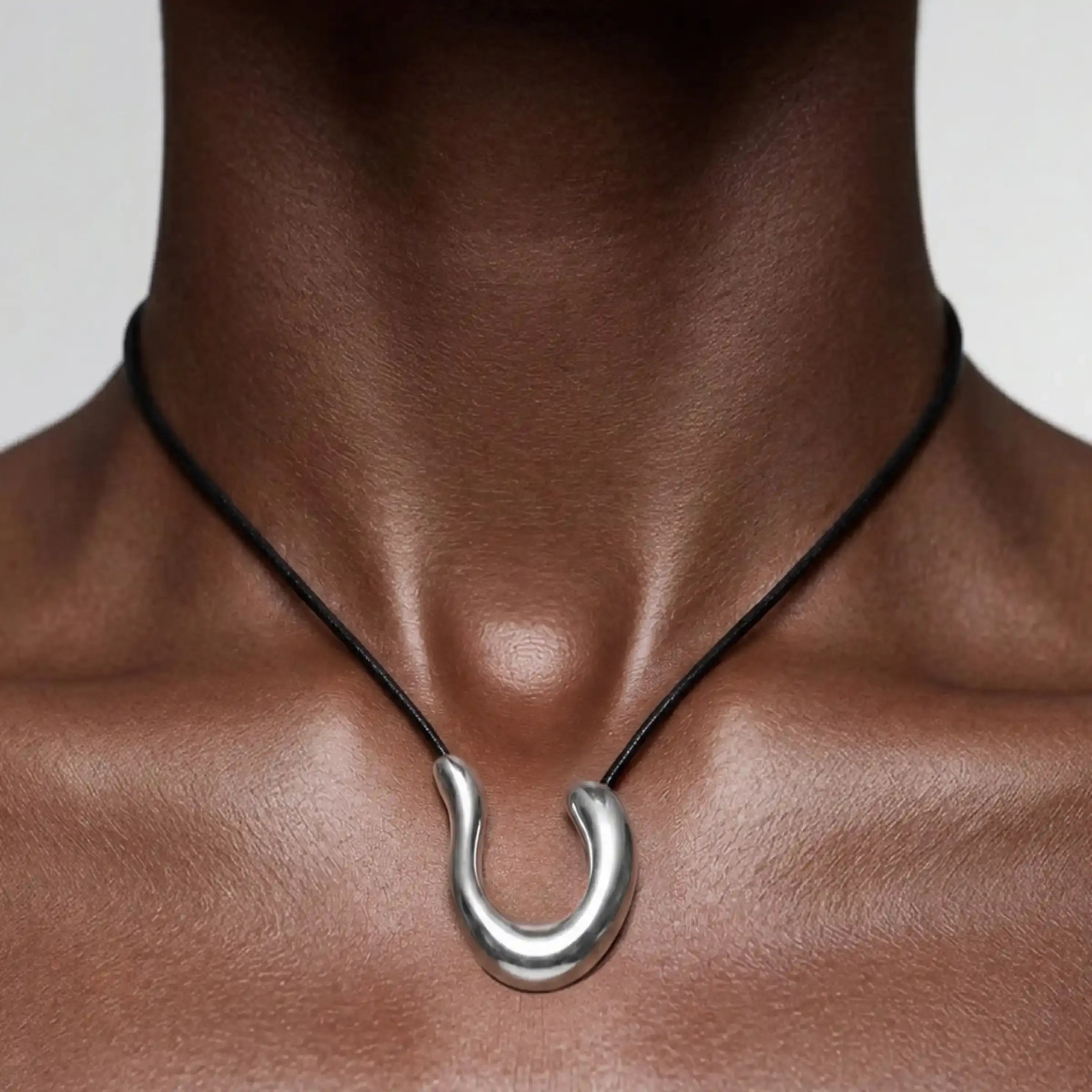 Pendentif en forme de fer à cheval argenté, suspendu à un cordon noir et porté au niveau de la clavicule.