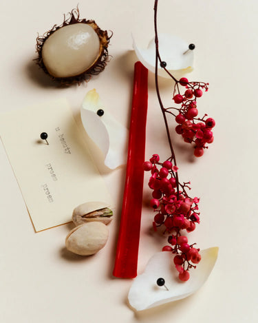 Flacon de parfum blanc avec une bougie rouge, entouré de ramboutans, de pistaches, de baies rouges et d'une fiche produit sur fond neutre