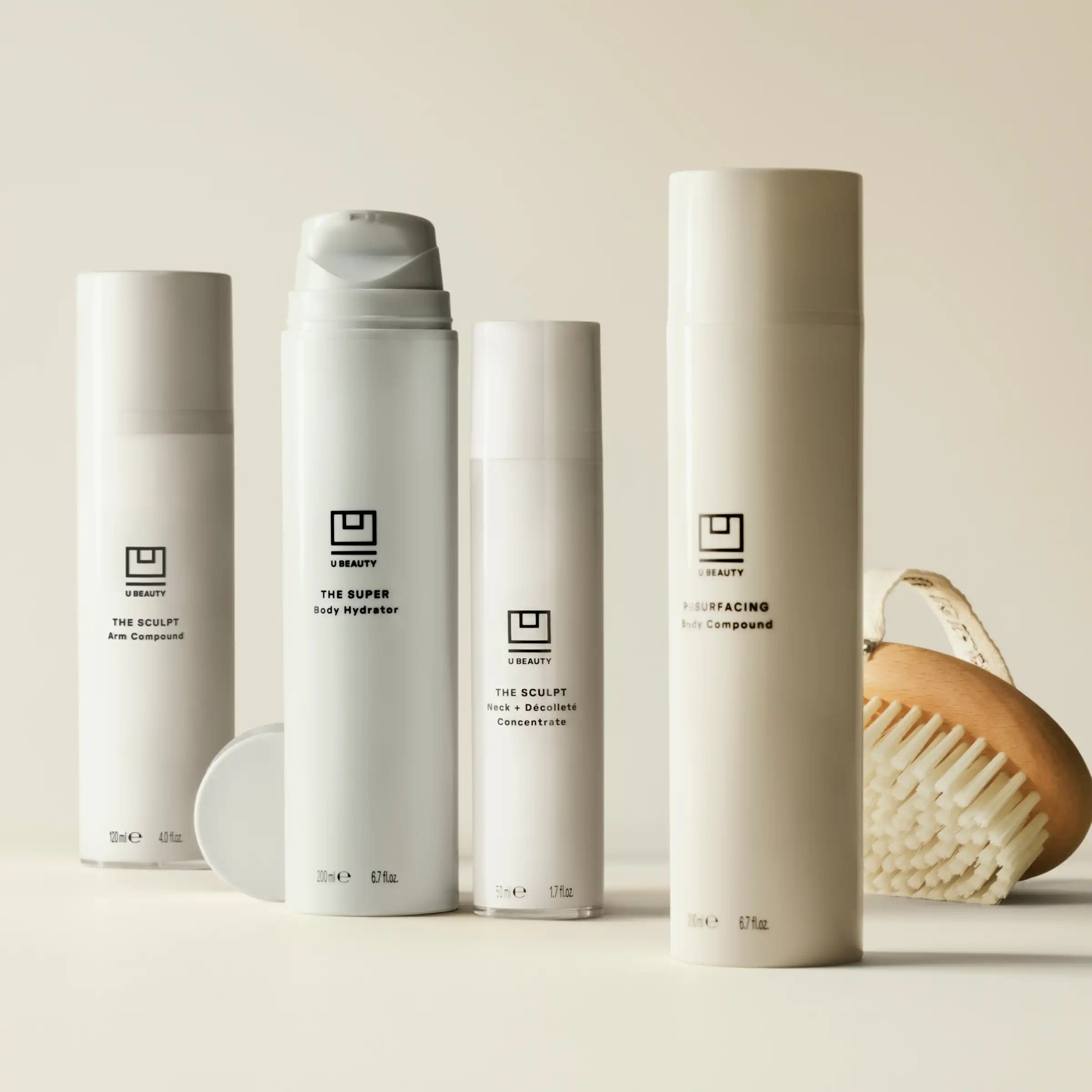 White cylindrical bottles from U Beauty skincare line arranged with a wooden brush on neutral background.
