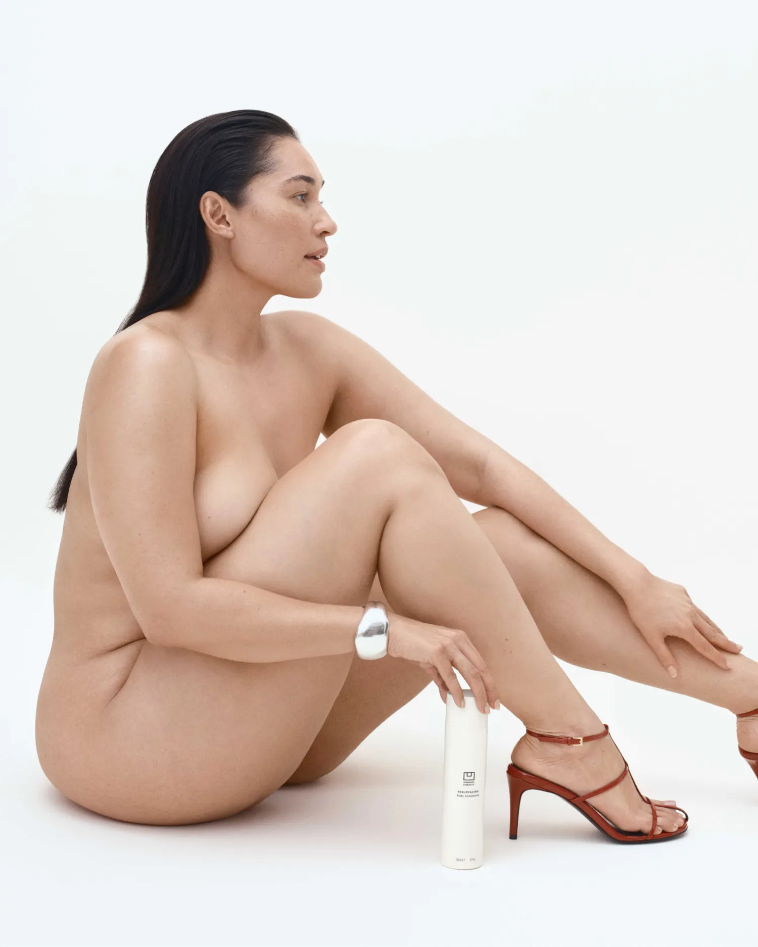 Une femme assise à côté d'un flacon cylindrique blanc orné d'une bande argentée, chaussée de sandales à talons rouges.