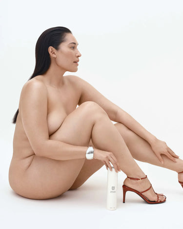 Une femme assise à côté d'un flacon cylindrique blanc orné d'une bande argentée, chaussée de sandales à talons rouges.