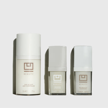 Three white cylindrical cosmetic bottles with taupe logos labeled "The Sculpt Arm Compound," "Eye Serum Hydrator," and "Resur